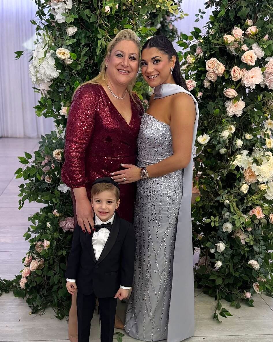 A mother and daughter with a little boy in a tuxedo in front of a floral wall